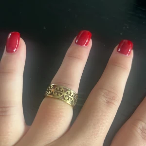 Guldfärgad ring från Tory Burch - Snygg guldfärgad ring från Tory Burch med det klassiska logomönstret runt hela ringen. Perfekt statement piece som lyfter din stil och passar till alla tillfällen. Ringen har en bred form och glänsande finish. Storlek på ringen 16/17
