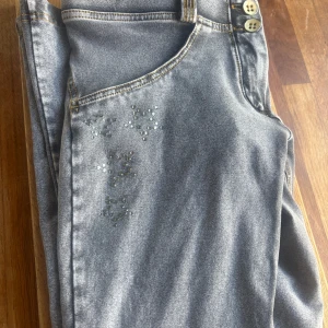 Grå WR.UP byxor med strassdetaljer - Snygga grå byxor från Freddy WR.UP med coola strassdetaljer på fickor och framsida. Byxorna har två knappar i midjan, kontrastsömmar och en figurnära passform. Perfekta för dig som vill ha något extra till din outfit.