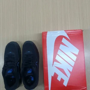 Nike Air Max svarta/blå sneakers - Säljer ett par svarta Nike Air Max sneakers med blå detaljer och logga. Skorna har klassisk Air Max-sula, snörning och är tillverkade i en mix av syntet och textil. Perfekta för dig som gillar sportig och stilren look.