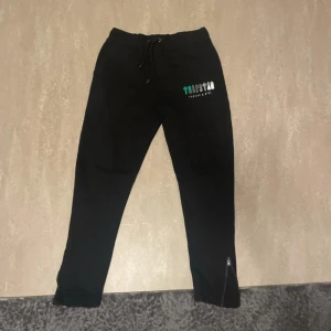 Svarta mjukisbyxor från Trapstar M - Svarta mjukisbyxor från Trapstar med logga på vänster höft. Byxorna har snörning i midjan och dragkedjor vid bensluten. Sköna och stilrena, perfekta för chill eller streetwear. Tillverkade i mjukt material.