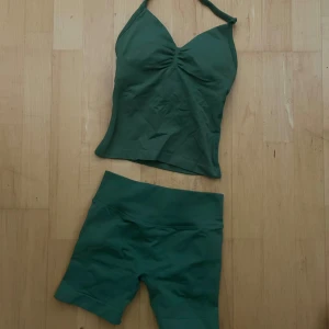 Grönt träningsset med hög midja och topp - Säljer ett grönt träningsset med högmidjade shorts och matchande topp med halterneck och rynkade detaljer både fram och bak. Båda delarna är sömlösa och stretchiga, vilket ger en bekväm passform. Perfekt för dig som vill ha en sportig och trendig look på stranden. Upplevs liten i storleken - passar trodligen någon med storlek XXS