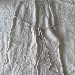 Beige omlottkjol i linne - Snygg beige långkjol i omlottmodell med knytband i midjan. Kjolen är tillverkad i linne och har en naturlig, avslappnad look. Perfekt för dig som gillar en enkel och stilren vibe. Den har en slits på sidan som en extra deltalj på kjolen. Den har tyvärr gått upp lite vid slitsen men inget som man tänker på.
