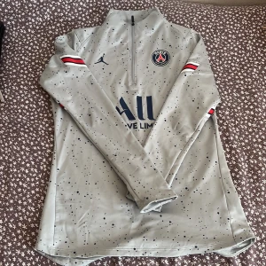 Grå långärmad PSG fotbollströja - Snygg grå långärmad PSG fotbollströja från Jordan med svart prickigt mönster och halv dragkedja. Tröjan har PSG-logga på bröstet, Jordan-logga och tryck i marinblått. Röd, vit och svart rand på ärmarna. Perfekt för träning eller match.