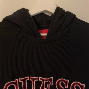 Svart hoodie från Guess med broderi - Svart hoodie från Guess i storlek M med stort broderat GUESS-tryck i vitt och rött på bröstet. Tröjan har huva, ribbade detaljer på ärmar och huva samt en klassisk känguruficka framtill. Perfekt för chill dagar och streetstyle.