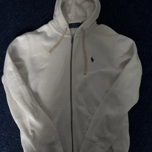 Vit hoodie från Polo Ralph Lauren - Säljer en stilren vit hoodie från Polo Ralph Lauren i bomull. Tröjan har dragkedja framtill, huva med snörning och klassisk logga broderad på bröstet. Perfekt för en chill och clean look. Ficka framtill för extra bekvämlighet.