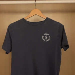 Mörkblå t-shirt från Polo Ralph Lauren - Snygg mörkblå t-shirt från Polo Ralph Lauren med klassisk logga tryckt på bröstet. Tillverkad i mjuk bomull som känns skön mot huden. Perfekt för en chill och stilren look. Storlek 14-16 skulle säga i storlek XS/S