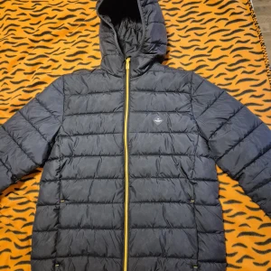 Svart dunjacka från Hackett London - Snygg svart dunjacka från Hackett London med huva och gul dragkedja. Jackan är quiltad och har en stilren look med diskret logga på bröstet. Perfekt för kalla dagar när du vill hålla stilen och värmen.