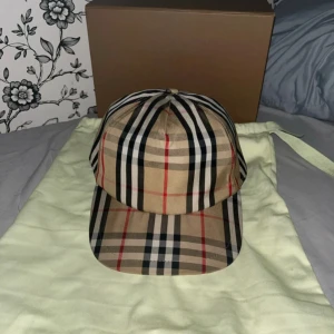 Burberry rutig keps i bomull - Säljer en klassisk keps från Burberry med det ikoniska rutiga mönstret i beige, svart, vitt och rött. Kepsen har justerbar baksida och tydlig Burberry-logga bak. Tillverkad i bomull och levereras med originalpåse och box. 10/10 skick