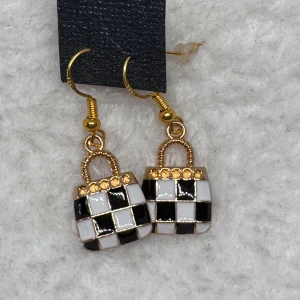 Svartvita rutiga örhängen med guld - Unika örhängen formade som små väskor med svartvitt rutmönster och guldfärgade detaljer. Ovanpå finns små glittrande stenar som ger extra bling. Perfekt accessoar för dig som gillar statement-smycken och cool design. Krokarna är i sterling silver 