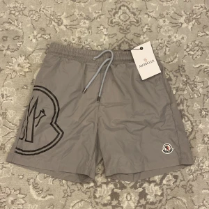 Grå Moncler shorts med snörning - Snygga grå shorts från Moncler med stor logga på vänster ben och klassisk patch framtill. Modellen har elastisk midja med snörning och bakficka. Perfekta för sommaren och chill dagar, tillverkade i lätt polyester som torkar snabbt. Perfekta som badshorts på sommaren eller som stilrena tränings-/casualshorts.