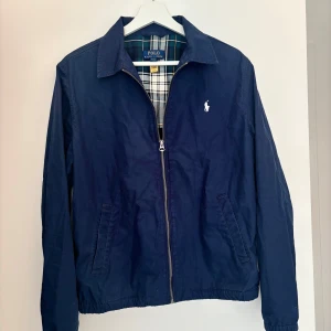 Marinblå vindjacka från Polo Ralph Lauren - Snygg marinblå vindjacka från Polo Ralph Lauren med klassisk krage och dragkedja framtill. Jackan har två fickor på sidorna och ett rutigt innerfoder i blått, grönt och gult. Perfekt för dig som vill ha en stilren och tidlös look.