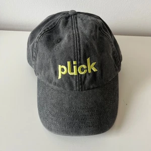 Grå keps från Plick med brodyr - Snygg grå keps från Plick med gul broderad logga framtill. Klassisk böjd skärm och justerbart spänne baktill i metall. Helt ny!