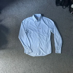 Ljusblå skjorta från Lexington - Klassisk ljusblå skjorta från Lexington med button-down krage och bröstficka. Skjortan har långa ärmar, knappar hela vägen och är tillverkad i mjuk bomull. Perfekt för en clean och stilren look.