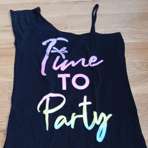 Svart oneshoulder klänning BluKids - Svart kortklänning från BluKids med oneshoulder-design och ett smalt axelband. Framsidan har färgglad text 'Time to Party' i pastellfärger. Mjuk och skön bomull, perfekt för dig som gillar en cool och avslappnad stil.