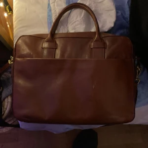 Brun datorväska i skinn från Bläck - Snygg brun datorväska från Bläck i äkta skinn. Klassisk design med två handtag, avtagbar axelrem och dragkedja upptill. Perfekt för laptop och dokument, med flera innerfack för smart organisering. Stilren och tidlös look som passar till allt.