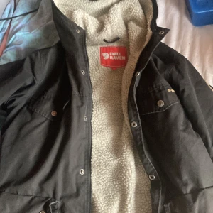 Svart teddyjacka från Fjällräven XS - Svart teddyjacka från Fjällräven med mysigt vitt teddyfoder och klassisk logga på ärmen. Jackan har tryckknappar, dragkedja och två bröstfickor. Perfekt för kalla dagar när du vill vara både varm och snygg.