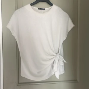 Vit topp med knyt från Zara - Säljer en stilren vit topp från Zara med korta ärmar och snygg knytdetalj i sidan. Toppen har en rund halsringning och är tillverkad i ett mjukt material som känns skönt mot huden. Perfekt för en enkel men trendig look.