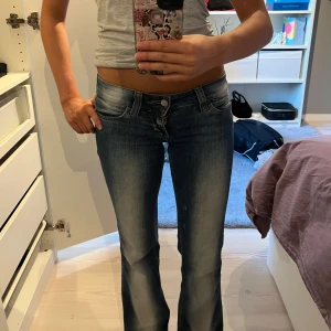 Blå bootcut jeans från Weekday - Säljer ett par blå bootcut jeans från Weekday med låg midja och snyggt vita detaljer. Jeansen har klassisk femficksdesign, dragkedja och knapp framtill. Materialet är mjukt denim i bomull och passformen är normal med utsvängda ben. Jeansen har ett hål längst ner på baksidan!💞