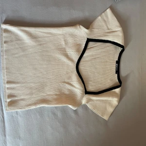 Ribbad vit topp med svart kant från Bikbok - Superfin ribbad topp i vit med svart kant runt urringningen från Bikbok. Toppen är croppad, har korta ärmar och en snygg, lite djupare fyrkantig ringning. Perfekt att styla med jeans eller kjol för en trendig look.