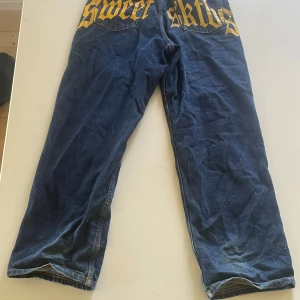 Sweet sktbs jeans - Säljer ett par blå jeans med rak passform och cool gul text i gotisk stil på bakfickorna. Jeansen har klassisk femficksdesign och är tillverkade i jeansmaterial. Perfekta för dig som vill sticka ut lite extra med din stil.