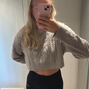 Beige croppad stickad tröja från Bershka - Säljer en beige croppad stickad tröja med snyggt kabelstickat mönster och bred ribbad kant nedtill. Tröjan har lång ärm och en relaxed fit som ger en chill vibe. Perfekt att styla med höga jeans eller kjol för en trendig look.