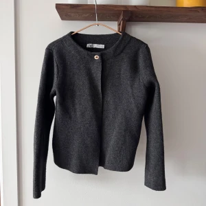 Mörkgrå kofta från Zara - Stilren mörkgrå kofta från Zara med rund halsringning och en enkel knapp framtill. Koftan är långärmad och har en clean look som passar perfekt till lager-på-lager. Materialet är mjukt ullblandning. Snygg på kontor/jobb/skola. 