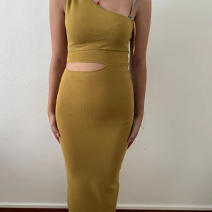 Gul one shoulder klänning med cut-out - Snygg gul långklänning med one shoulder och cut-out detalj vid midjan. Klänningen har en tight passform och är ärmlös, perfekt för dig som vill sticka ut. Materialet är stretchigt och följsamt.