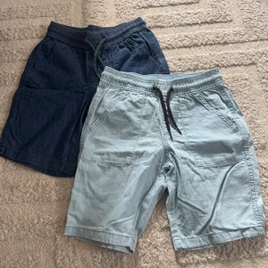 Två par shorts från Lupilu, blå & ljusblå - Säljer två par shorts från Lupilu i storlek 122/128. Ena paret är mörkblå och det andra ljusblå, båda med elastisk midja och snörning. De har praktiska fickor på sidorna och är gjorda i mjuk bomull, perfekta för chill dagar under sommaren.