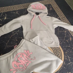  Syna world TrackSuit - Snygga grå Mjukis byxor med elastisk midja och rosa snörning. På ena benet finns ett stort rosa graffiti-inspirerat tryck som ger en cool streetkänsla. Mjukt material, perfekt för chill eller träning. Matcha med hoodie för en komplett look.
