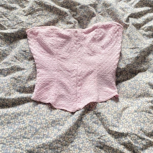 Ljusrosa korsett topp i strukturerat tyg - Supersöt ljusrosa korsett topp med strukturerat, vågigt tyg och markerade kupor. Axelbandslös bandeau-modell som ger en snygg siluett. Perfekt att styla med jeans eller kjol för en trendig look.