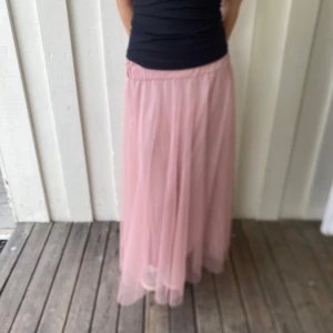 Rosa långkjol i tyll från Gemini - Supersöt rosa långkjol från Gemini i flera lager tyll som ger en drömmig och flowy känsla. Kjolen har resår i midjan för extra bekvämlighet och passar perfekt till en trendig och romantisk look. Perfekt för dig som vill sticka ut med en feminin vibe.