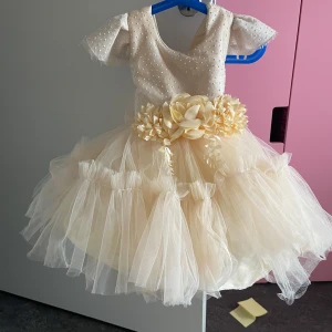 Champagnefärgad prinsessklänning med rosett - Superfin champagnefärgad prinsessklänning med puffiga korta ärmar, pärldetaljer på överdelen och stor blomma framtill i midjan. Kjolen är fluffig med lager av tyll och baktill finns en stor rosett. Perfekt för speciella tillfällen och ger en magisk känsla.passar upp till två år