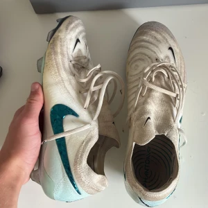 Nike Phantom GX Elite fotbollsskor vita - Nike Phantom GX Elite fotbollsskor i vitt med turkosblå Swoosh och cool struktur på ovandelen. Skorna har snörning och en genomskinlig, skimrande sula med dobbar för gräsplan. Lätta och smidiga för maxad bollkontroll och speed. Dom är användna 3 träningar har box o så till