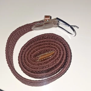 Brunt flätat bälte med skinn-detaljer - Snyggt brunt bälte med flätad design och detaljer i ljusbrunt skinn. Bältet har en silverfärgad metallspänne och vita sömmar på skinnpartierna. Perfekt accessoar för att lyfta din outfit med en klassisk touch.