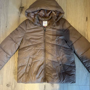 Brun pufferjacka från Vero Moda strl 164 - Säljer en brun pufferjacka från Vero Moda med huva och dragkedja framtill. Jackan har två fickor med knappstängning och är perfekt för kyliga dagar. Den har en enkel, stilren look och är lätt att matcha med olika outfits. 💗