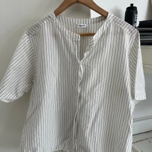 Vit randig henley från Filippa K - Vit henley-skjorta från Filippa K med tunna svarta vertikala ränder. Skjortan har korta ärmar, rak passform och en enkel krage utan knappar framtill. Tillverkad i mjuk bomull, perfekt för en clean och stilren look.