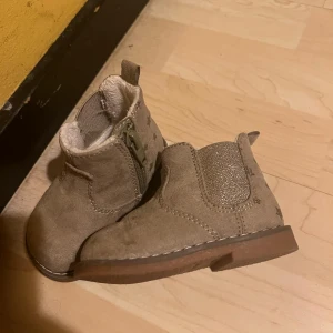 (Barnskor)Beige fodrade boots med stjärnor från H&M - (Barnskor)Beige fodrade boots från H&M i mockaimitation med glittrig resår på sidan och stjärnmönster över hela skon. Fodrade och har dragkedja på sidan för enkel påtagning. Storlek 18-19 