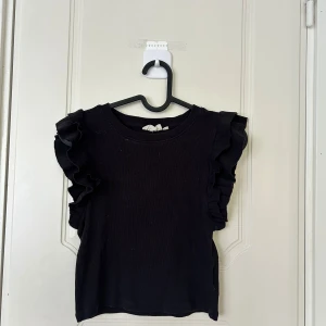 Svart volangtopp från UNIQ Studio - Svart ärmlös topp från UNIQ Studio med breda volangdetaljer längs axlarna. Toppen är ribbad och har en rund halsringning. Perfekt för dig som gillar en enkel men ändå cool look med extra edge.