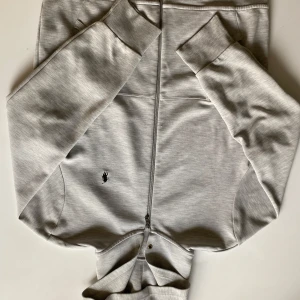 Grå Polo Ralph Lauren zip-hoodie - Säljer en ljusgrå Polo Ralph Lauren zip hoodie i storlek M. Använd mycket sparsamt och ser nästan ny ut, men tyvärr lite för stor för mig. ✅ Stilren design utan snören ✅ Klassiska loggan på bröstet ✅ Mycket bra skick, nästan ny ⚠️ Liten blå prick på vänster underarm ⚠️ Två små hål vid händerna (går lätt att fixa) 💰 Pris kan diskuteras