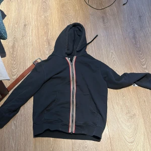 Burberry hoodie - Säljer en burberry hoodie med dragkedja och randiga detaljer längs dragkedjan. Dragkedjan saknar saken man drar i men inget som gör så mycket. Tröjan har en klassisk design med huva och fickor framtill. Perfekt för en avslappnad stil. Köpt på nk
