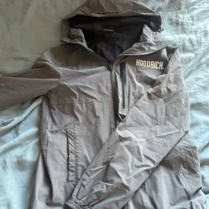 Grå vindjacka från Hoodrich - Säljer en grå vindjacka från Hoodrich med huva och dragkedja. Jackan har en bröstficka med dragkedja och elastiska ärmslut. Perfekt för blåsiga dagar!