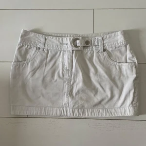 Vit kortkjol med bälte - Snygg vit kortkjol med bälte och fickor fram och bak. Perfekt för en somrig look. Kjolen har en stilren design med knappdetalj i midjan.
