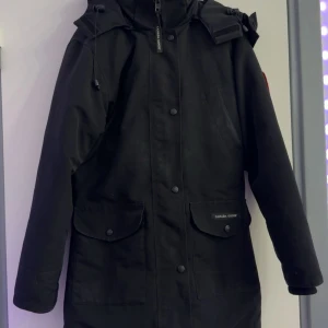 Svart parkajacka från Canada Goose - Säljer en äkta svart parkajacka från Canada Goose med lång ärm och dragkedja. Jackan har en klassisk design med två stora fickor framtill och en logotyp på ärmen. Perfekt för kalla dagar. Pris kan diskuteras undrar du något om jackan så skriv