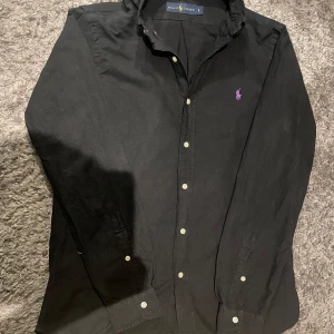 Svart skjorta från Ralph Lauren - Säljer en stilren svart skjorta från Ralph Lauren med klassisk krage och knappar framtill. Skjortan har en broderad lila logotyp på bröstet och långa ärmar. Perfekt för en elegant look.