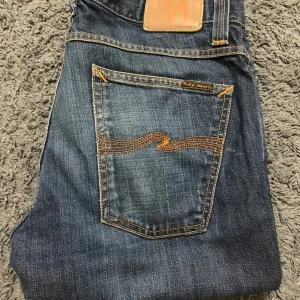  Nudie Jeans - Slim Jim - Säljer ett par klassiska blå jeans från Nudie Jeans med orange sömmar och en snygg broderad detalj på bakfickorna. Jeansen har en normal passform och är perfekta för en avslappnad stil. Ny pris -1800 kr. Modell Slim Jim