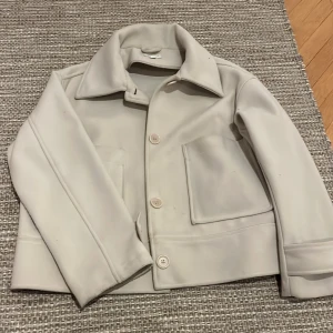 Vit kappa - Snygg vit kappa med stora knappar och klassisk krage. Jackan har en stilren design med två stora fickor framtill. Perfekt för en stilren look.