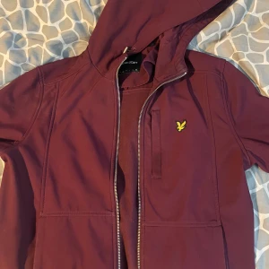 Vinröd jacka med huva från Lyle & Scott - Säljer en vinröd jacka med huva från Lyle & Scott. Jackan har en dragkedja framtill och en liten gul logotyp på bröstet. Perfekt för höst och vår med sin stilrena design och praktiska fickor.
