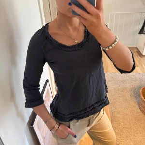 Svart topp med volangdetaljer - Säljer en svart topp från House med volangdetaljer vid halsringning och ärmslut. Toppen har trekvartsärmar och en bekväm passform. Perfekt för en stilren look.