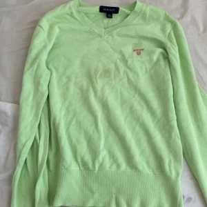 Grön tröja från Gant - Säljer en ljusgrön tröja från Gant med v-ringning och broderad logga på bröstet. Tröjan har långa ärmar och ribbade muddar vid ärmslut och nederkant. Perfekt för en stilren look!