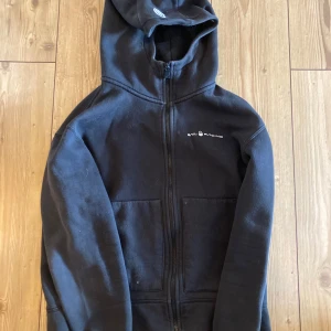 Svart huvtröja från Sail Racing - Säljer en svart huvtröja från Sail Racing med dragkedja och två fickor framtill. Perfekt för en avslappnad stil. Huvan ger extra värme och komfort. Ställ gärna frågor🌟